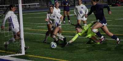 TMU women's soccer goalkeeper lunges for a soccer ball in front of the net, surrounded by TMU players and a U of T attacker
