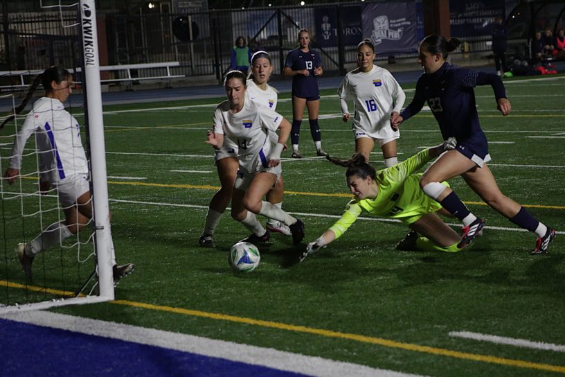 TMU women's soccer goalkeeper lunges for a soccer ball in front of the net, surrounded by TMU players and a U of T attacker