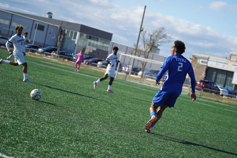 TMU soccer player approaches the ball against two Carleton defenders
