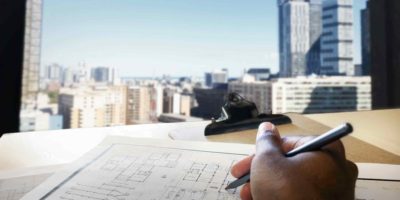 Photo of a someone holding a pencil over a desk filled with papers, looking out on a view of various buildings.