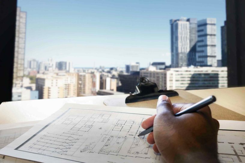 Photo of a someone holding a pencil over a desk filled with papers, looking out on a view of various buildings.