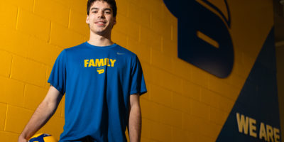 Photo of Riley Donovan in front of a yellow and blue wall with the TMU Bold logo, and "WE ARE BOLD" written on the wall.