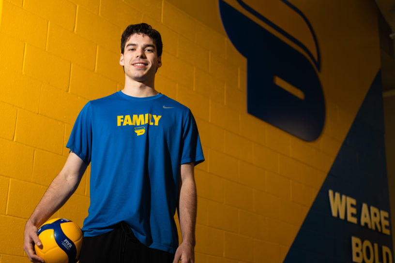 Photo of Riley Donovan in front of a yellow and blue wall with the TMU Bold logo, and "WE ARE BOLD" written on the wall.
